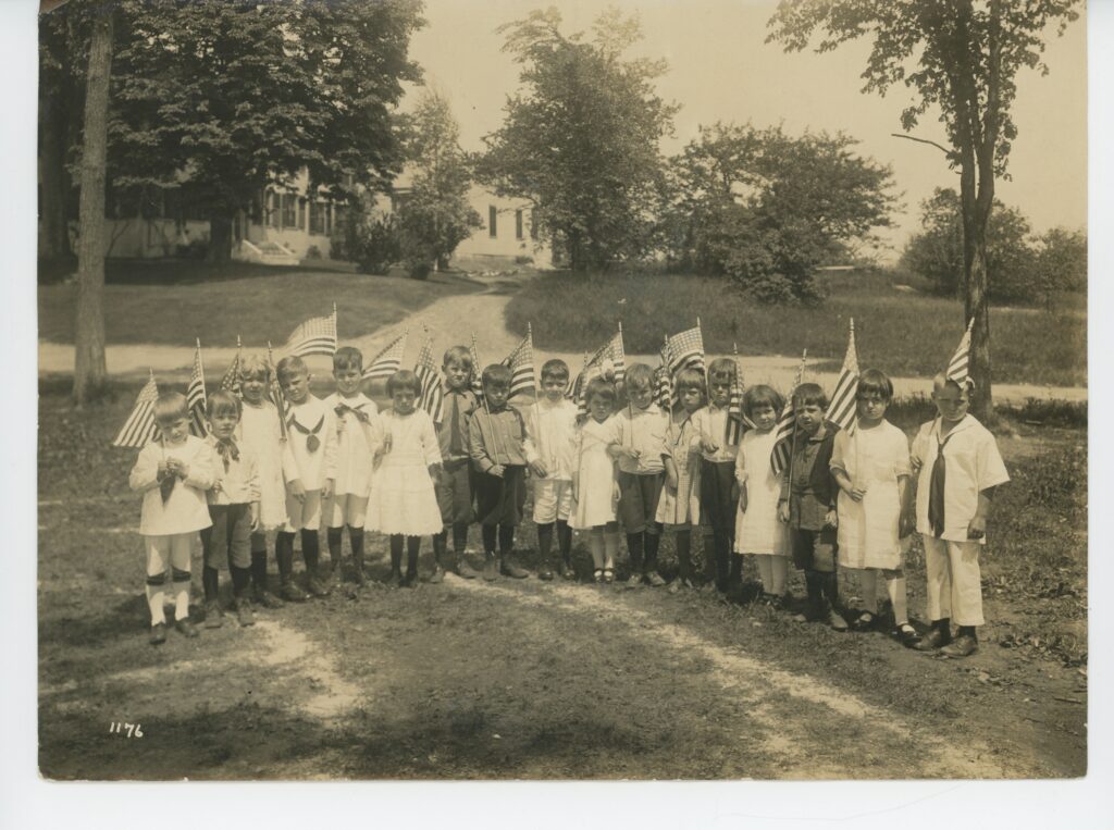 Black-and-white photograph of about twenty young children standing outdoors in a semicircle holding American flags, with a house and trees in the background; the child on the far right is identified as Raymond Lewis Swasey, circa 1919–1921.
