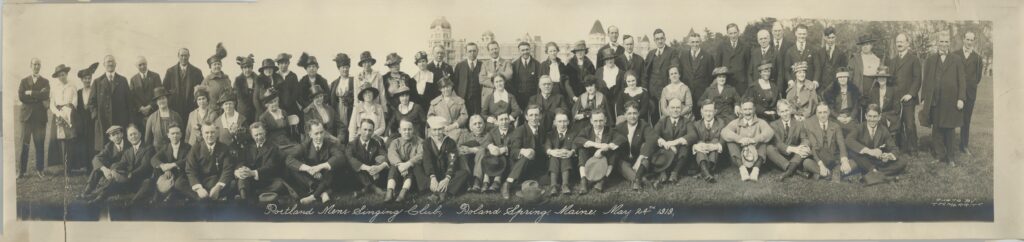 Panoramic group photograph of the Portland Men’s Singing Club and guests at Poland Spring House in Poland Spring, Maine, May 24, 1919, showing approximately seventy men and women arranged in three rows on the lawn in front of the resort hotel.