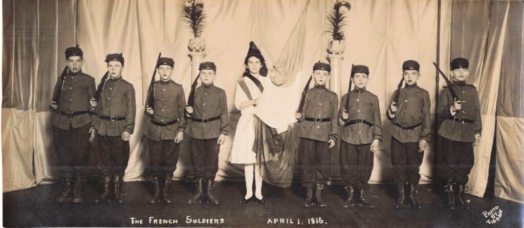 Group of eight Portland boys dressed as French soldiers with rifles, standing in formation on stage. A girl portraying France holding a French tricolor flag is center stage at the Burnham Gymnasium, Portland, Maine, April 1, 1916.