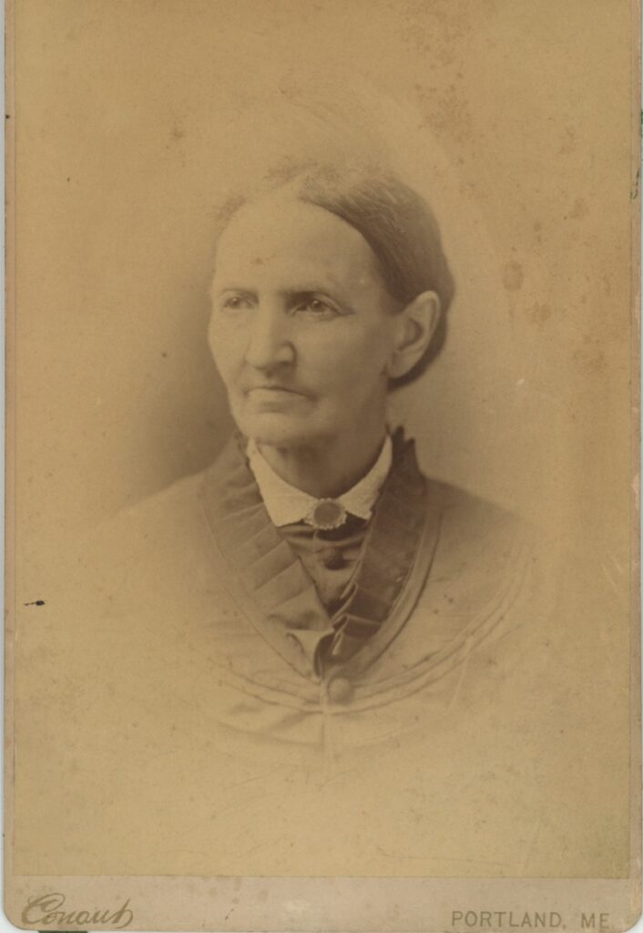 Cabinet card bust portrait of an older woman with dark hair pulled back, wearing a dark dress with a brooch, photographer Conaut, Portland, Maine, circa 1876–1884.