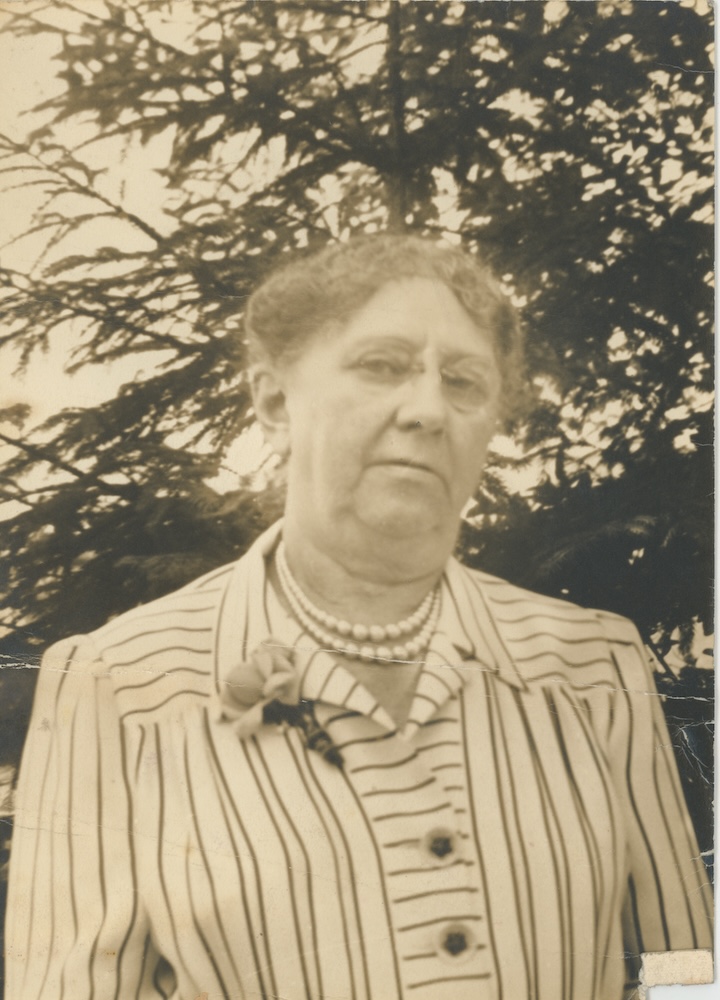 Sepia-toned portrait photograph of Eleanor Hinkley Kinney, a woman in her late fifties to mid-sixties, wearing a striped jacket, pearl necklace, and brooch, photographed outdoors with trees in the background.