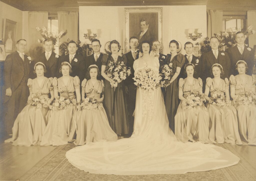 Formal sepia-toned wedding party portrait of approximately twenty people posed in an elegantly furnished private home, with the bride in a full-length white satin gown and cathedral train at center, surrounded by bridesmaids in light satin gowns and groomsmen in dark suits.