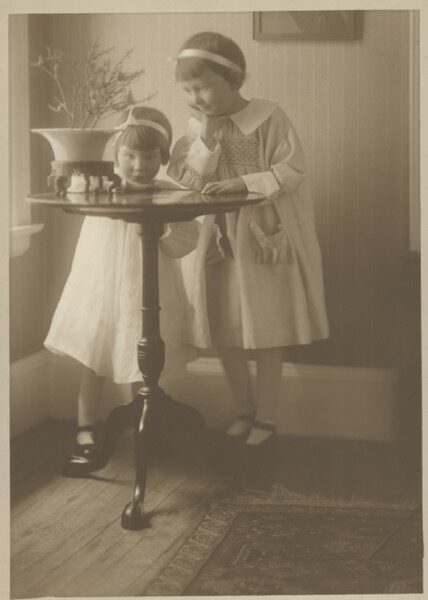 Sepia-toned photograph of two young sisters, Louise and Margaret Kinney, standing at a pedestal table in a home interior, circa 1917 to 1918.