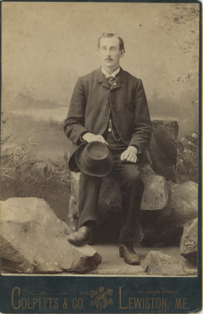 Cabinet card full-length portrait of a young man in a dark suit seated on a stone prop holding a bowler hat, photographer Colpitts and Co., Lewiston, Maine, circa 1890.