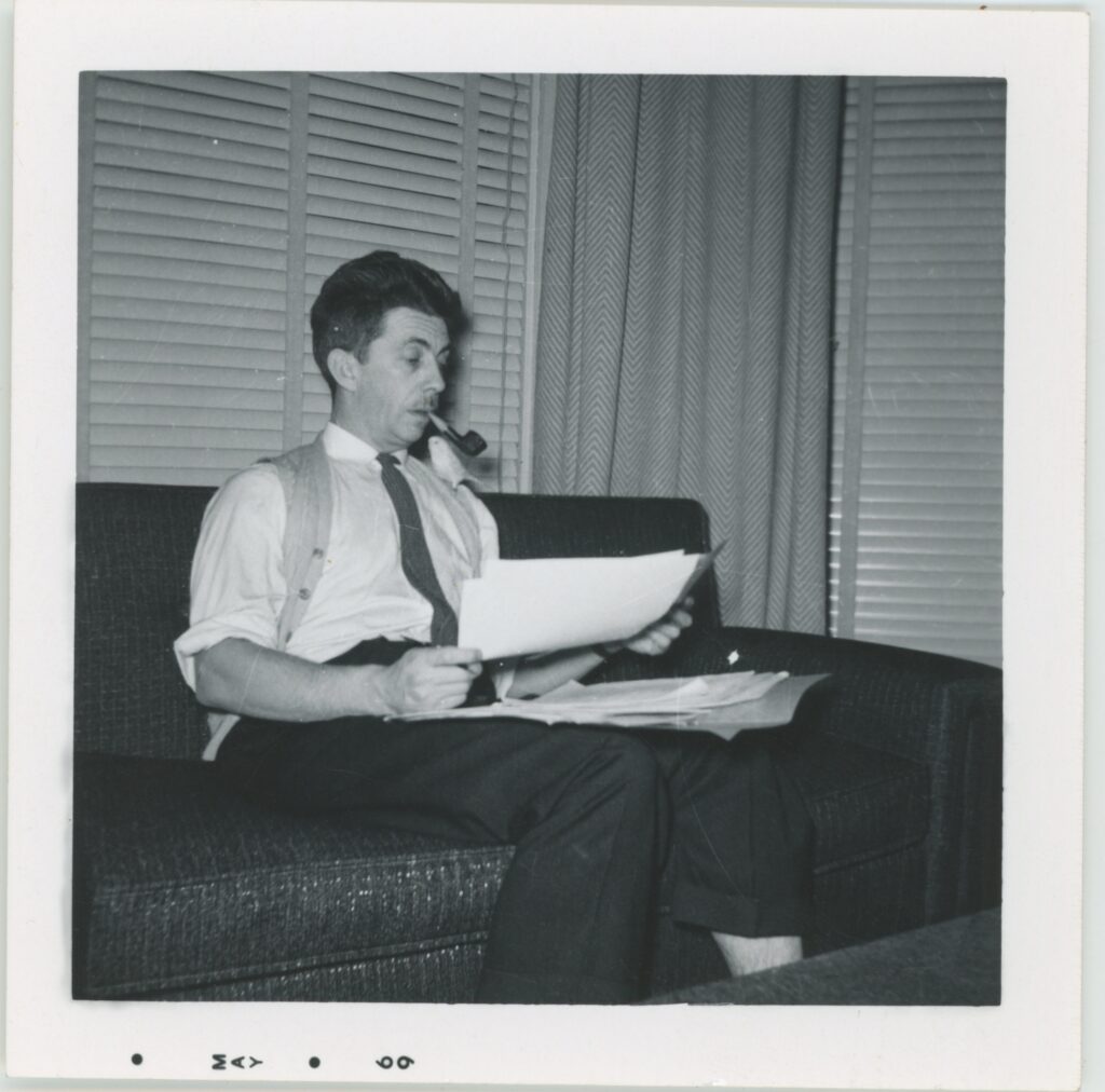 Man in vest and tie smoking a pipe, seated on a couch reading papers, 1950s.
