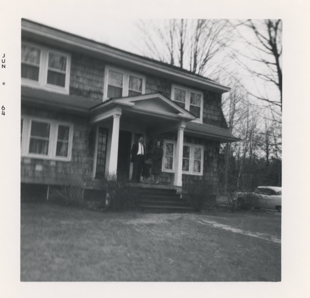 Two-story shingled house with covered porch, two people standing at the entrance, 1960s.