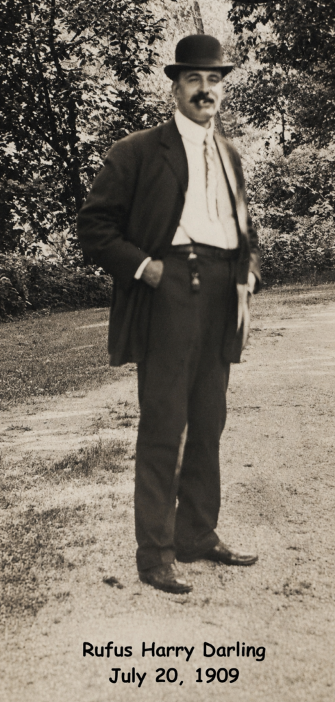 Man in dark suit and bowler hat standing outdoors on a dirt road, 1909.
