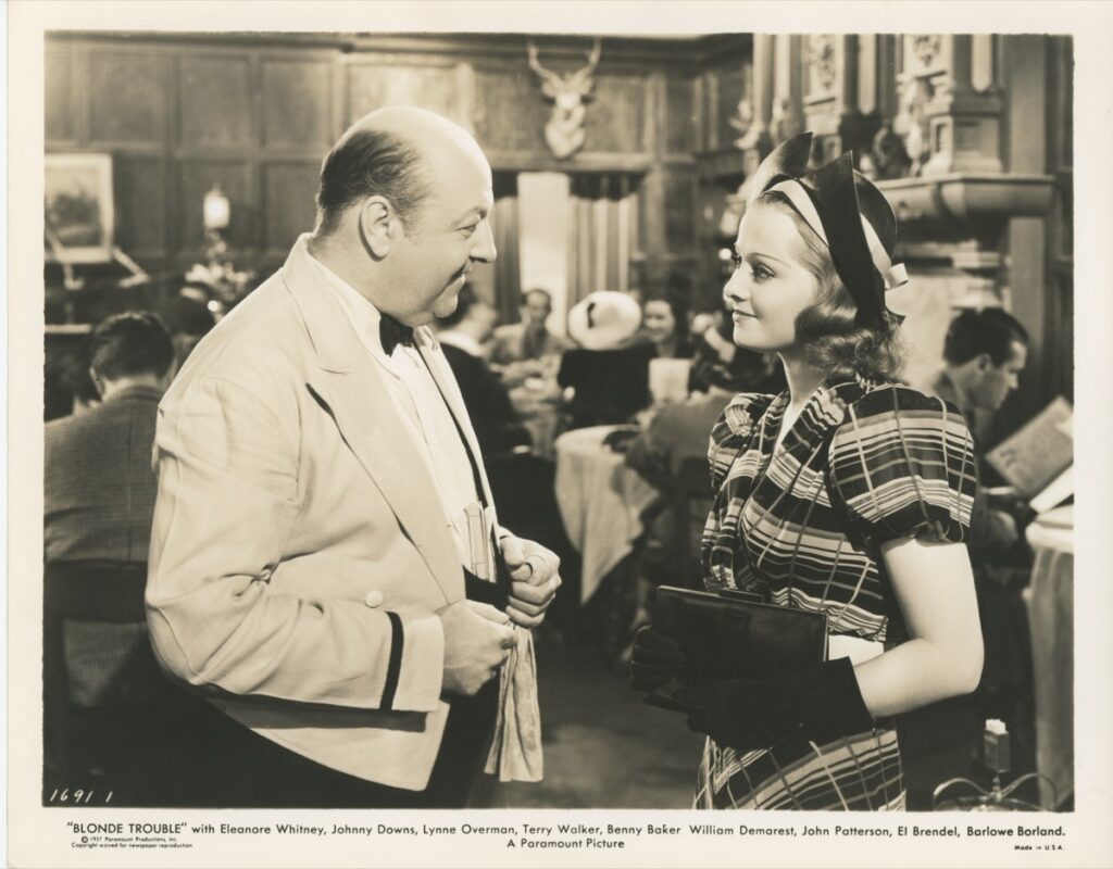 Photo — a young woman in a plaid dress and bow-trimmed hat greeted by an ol