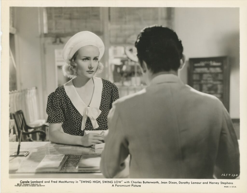 Photo — a woman in a polka-dot dress and sailor cap in conversation with a man, in an office setting with birdcages
