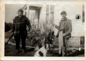 Photo of two men with guns, a string of pheasants, and a dog.