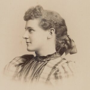 Photo of a young woman wearing a dress typical of the early 1890s. It is high-necked with a standing collar and large puffed sleeves. 