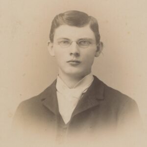 This is a portrait of a young man wearing a dark suit jacket and a high-collared white shirt. His large cravat and hairstyle are typical of the 1890s