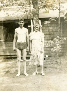 Photo of a young man in a 1930s swim suit and a woman standing by a cabin/camp. 