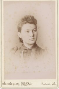 Sepia cabinet card portrait of a young woman named Maude Libby, approximately 16 to 22 years old, with dark wavy hair pinned up, wearing a high-collared pintucked blouse with a brooch and small drop earrings, photographed by Jackson of Portland, Maine, between 1891 and 1908.